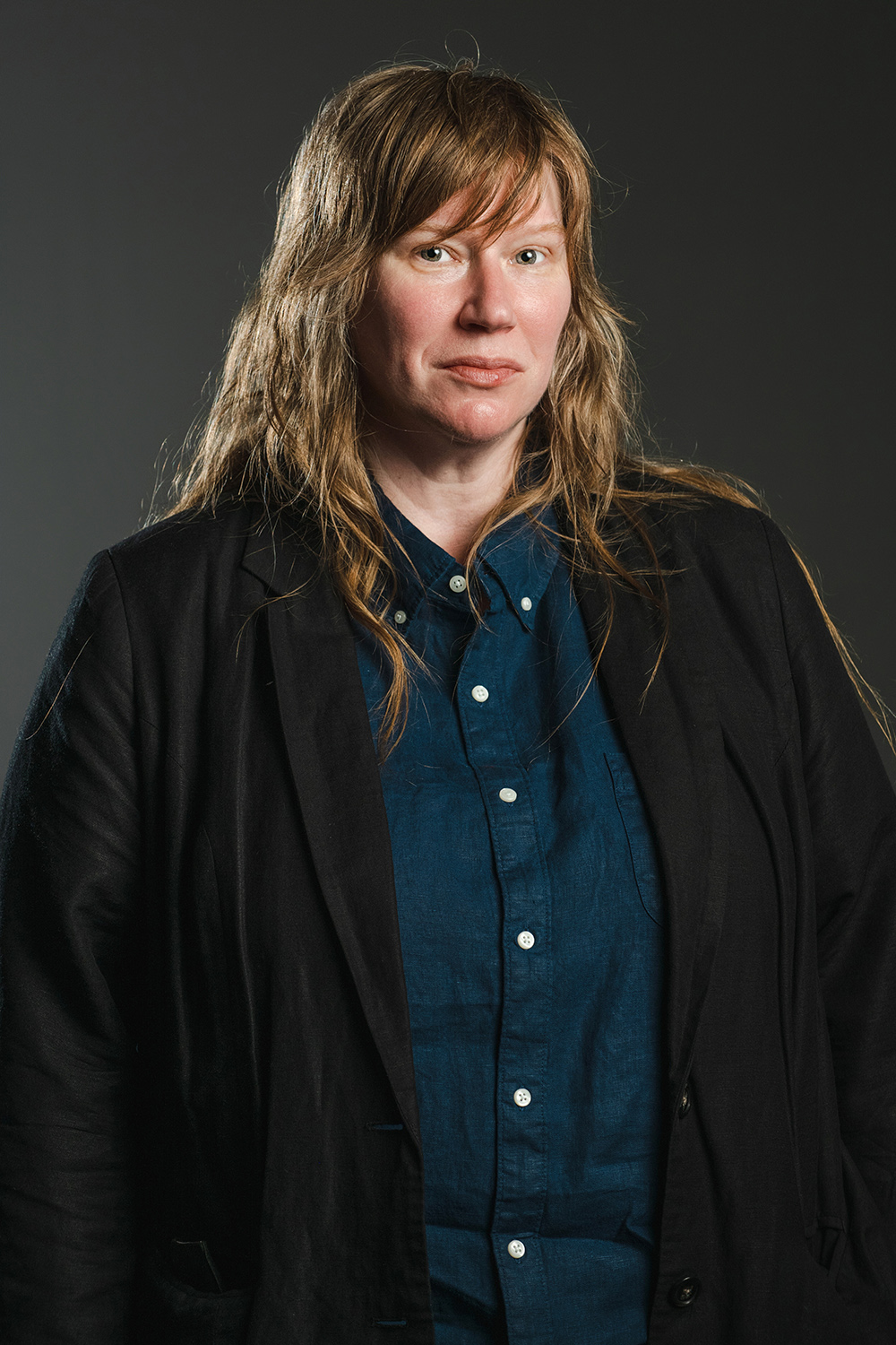 Color photograph of a light skinned woman with brown hair wearing a black blazer over an indigo shirt; she looks at the camera with a serious expression