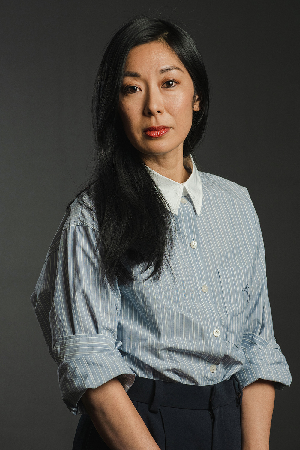 Color photograph of an Asian woman with long dark hair and a blue striped button up top with sleeves rolled, looking directly at the camera with a serious expression