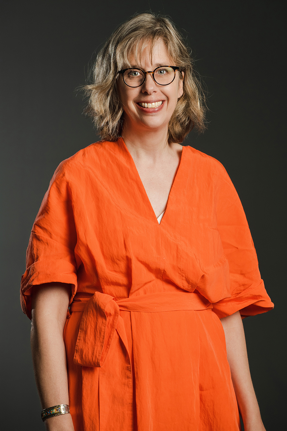 Color photograph of a light skinned woman with wavy blond hair wearing glasses and an orange jumpsuit