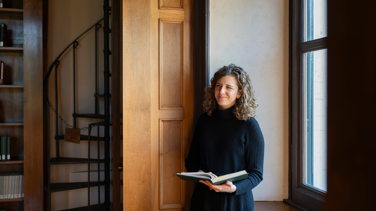 Color photo of a light skinned woman standing in a library holding an open book in her hands as she smiles at the camera