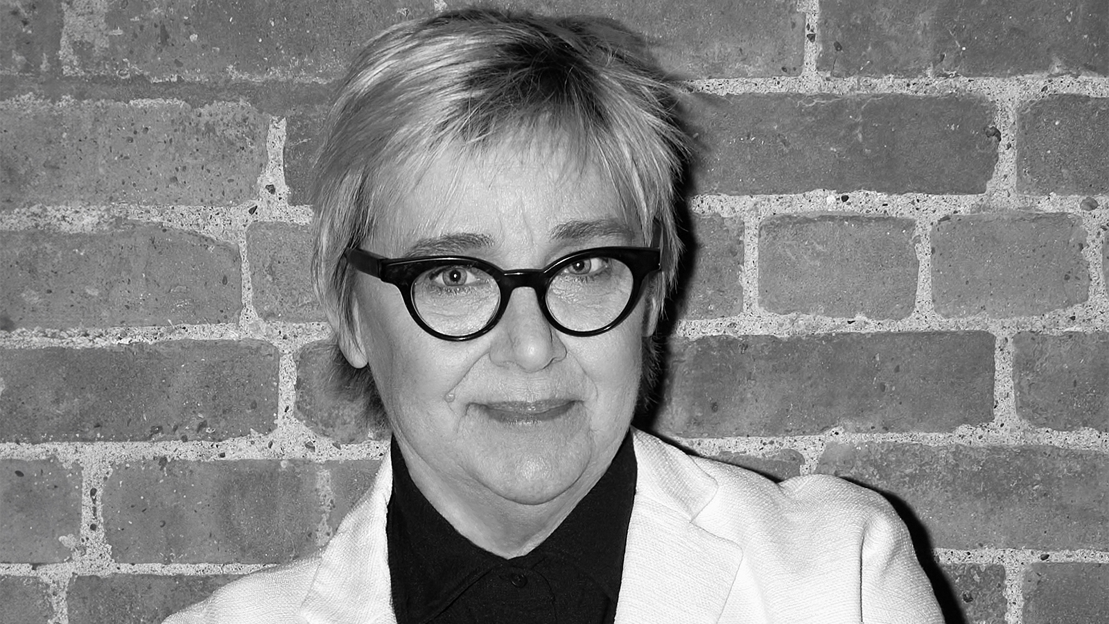 Black and white portrait of a light skinned woman wearing glasses and looking at the camera in front of a brick wall