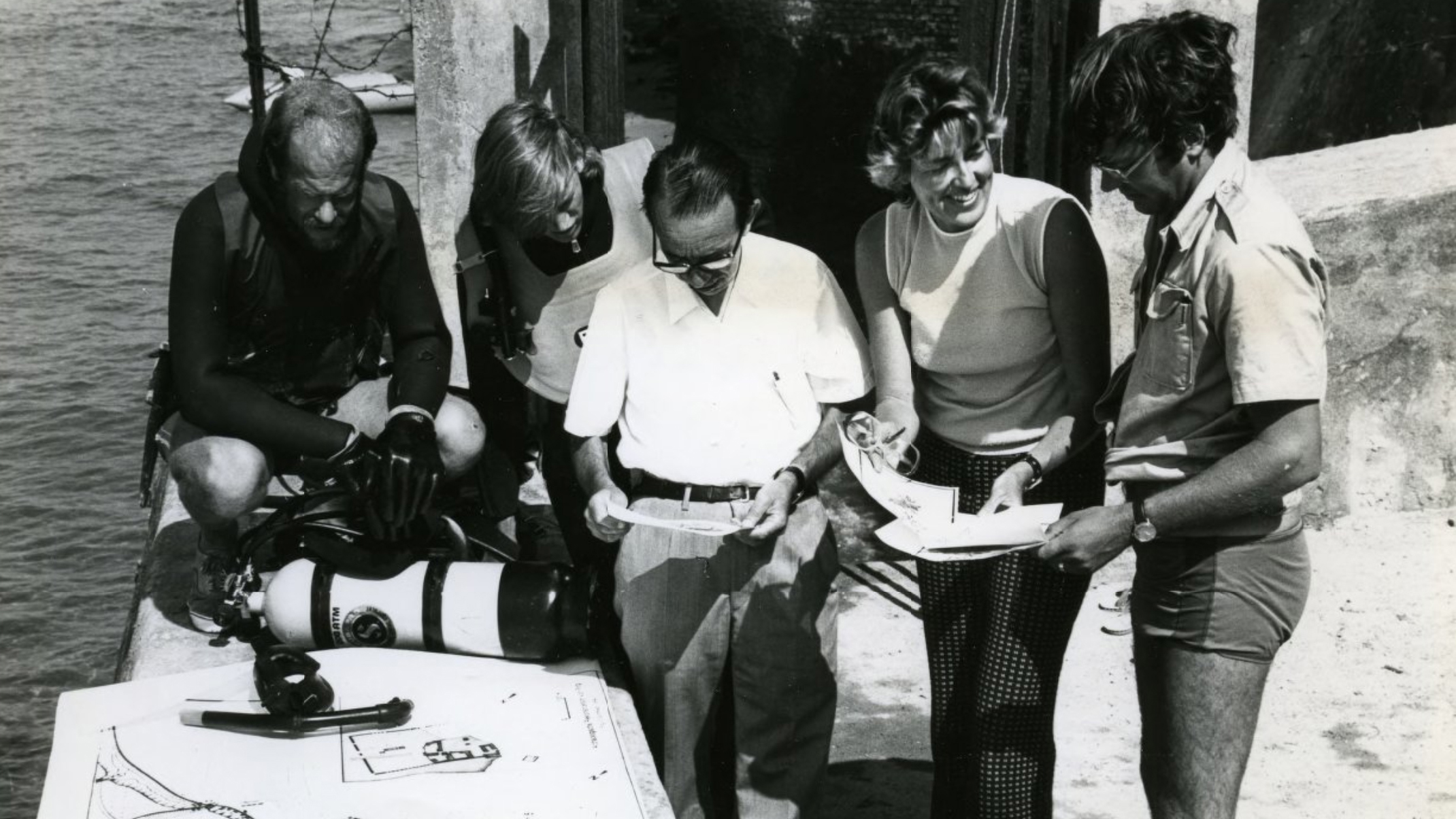 Black and white photograph of five people examining paper documents; SCUBA gear and water is visible to the left