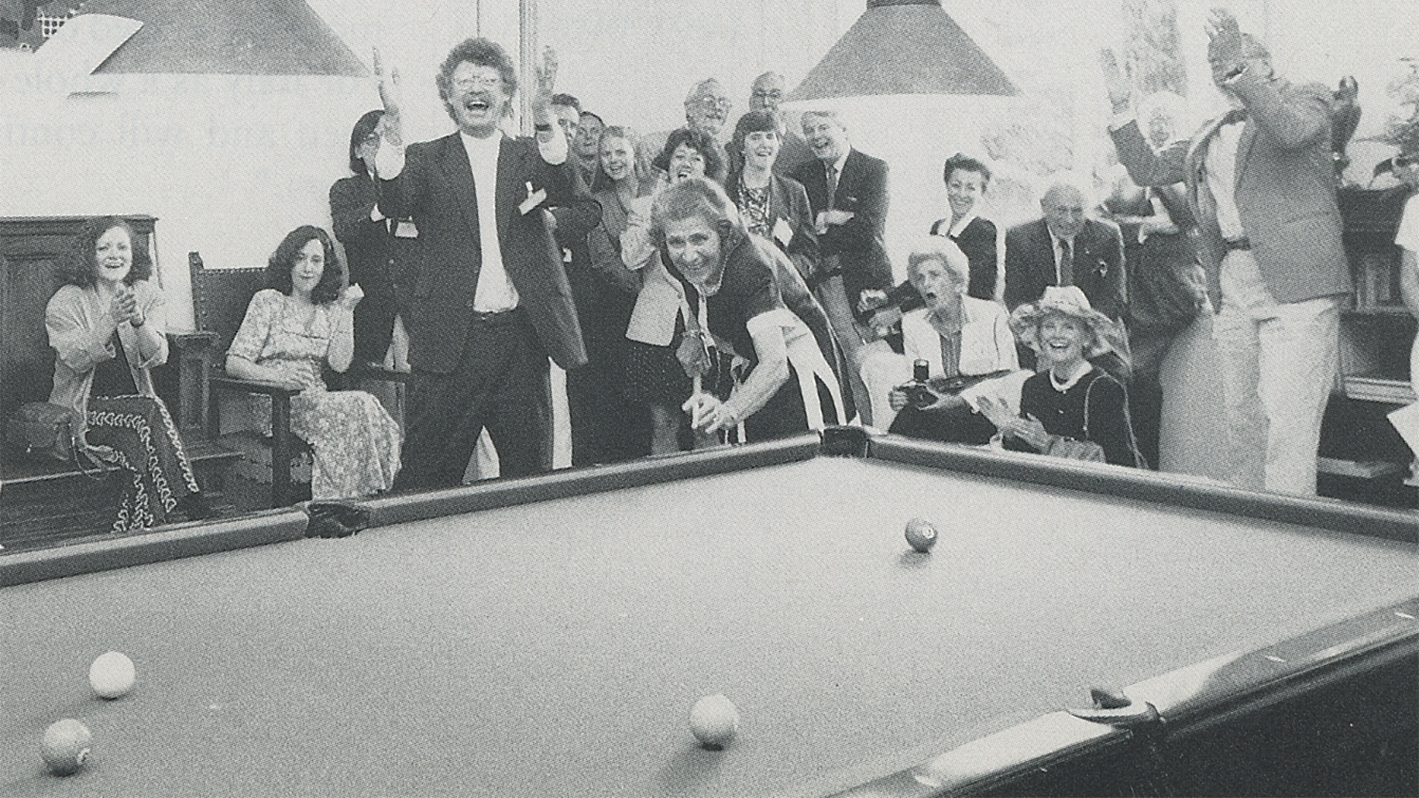 Black and white photograph from 1994 of many light skinned people clapping and cheering on a woman shooting pool in a salon