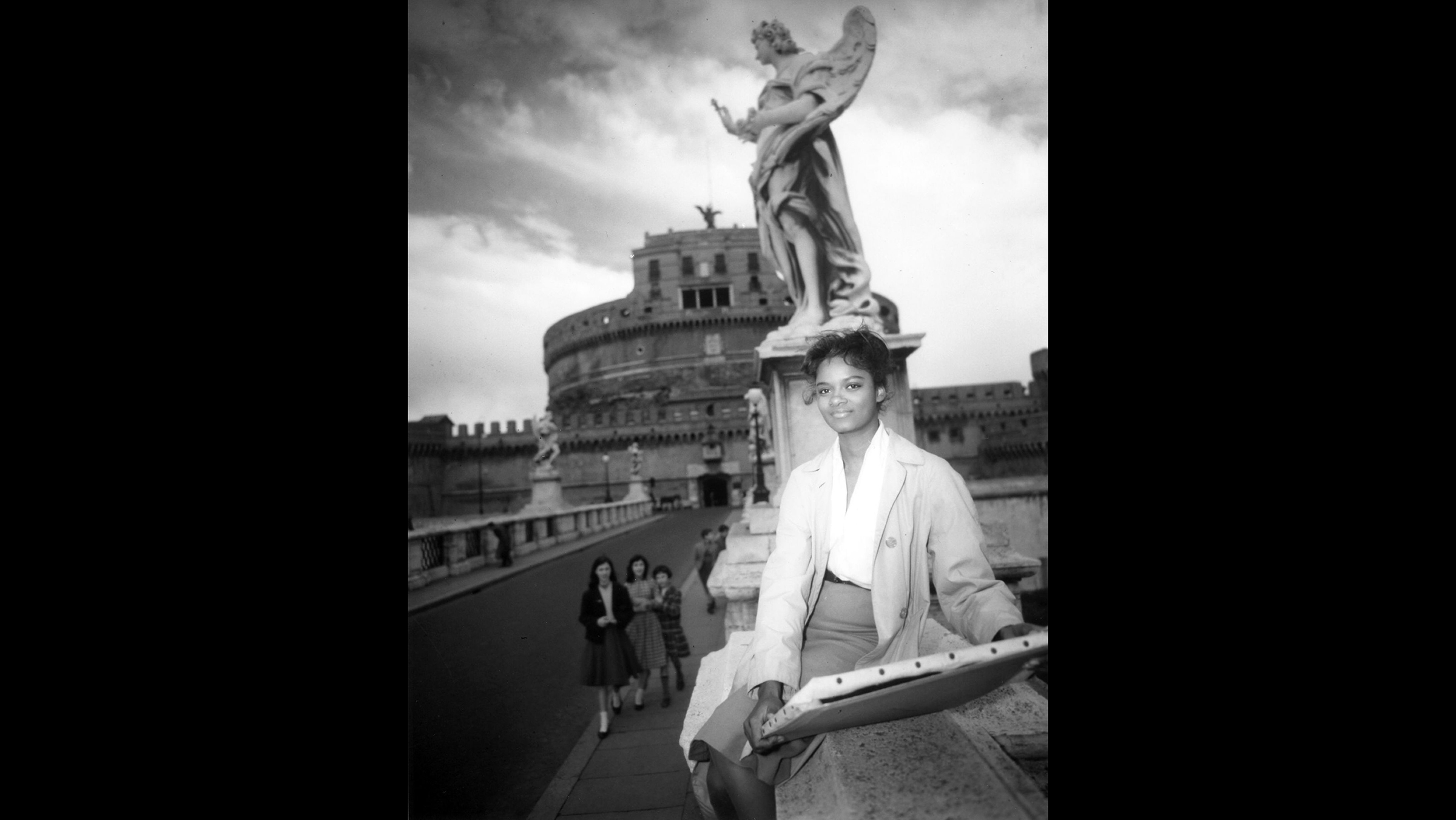 Black and white photograph of a young dark skinned woman sitting on the bridge in front of the Castel Sant'Angelo, an ancient Roman mausoleum; she holds a stretched painting canvas in her hand and stares into the distance as three young Italian girls on the bridge look up at her