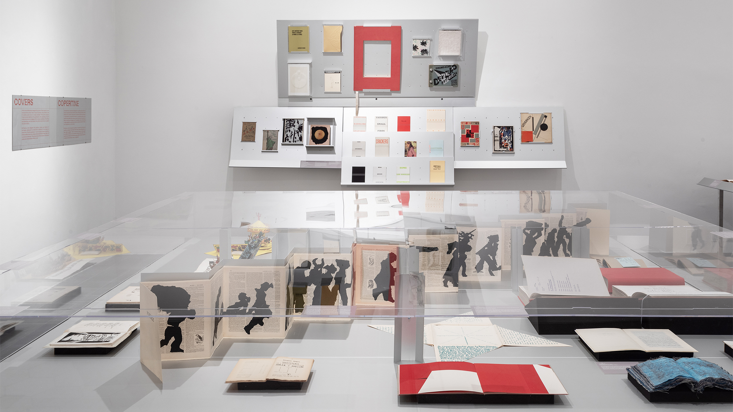 Color photo of an indoor art exhibition of artist's books, with publications under a large square plexicase and on wall shelves