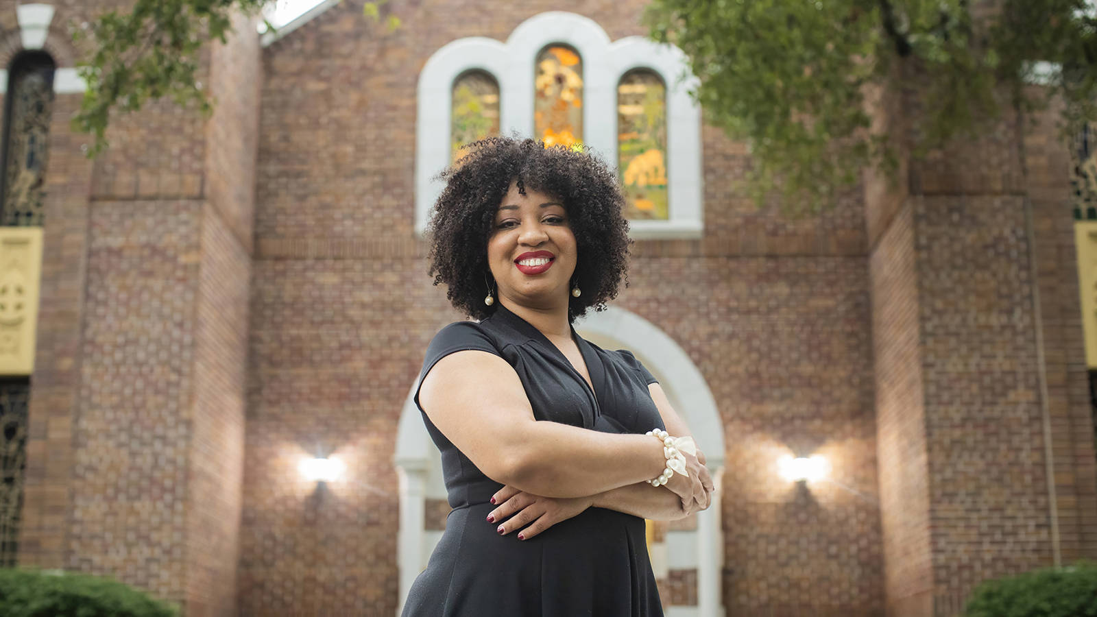 Color photograph of a dark skinned woman standing in front of a brick chapel; she wears a black dress, crosses her arms, and smiles at the camera