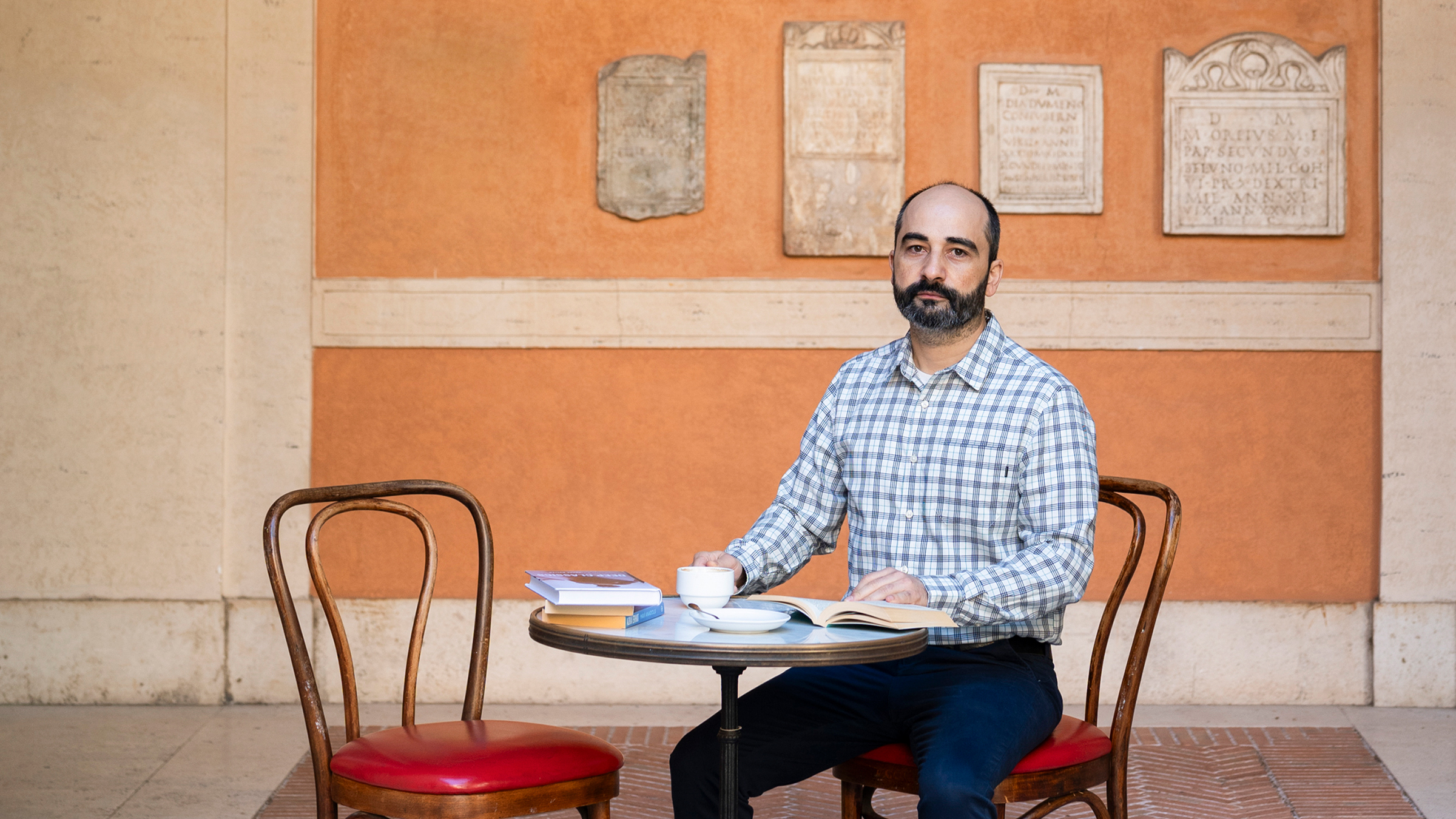 Color photo of an olive skinned man sitting at a cafe table with an open book, looking at the camera