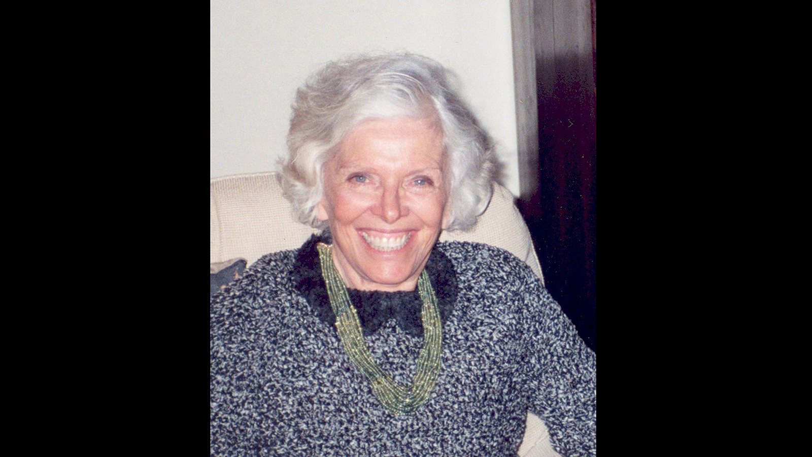 Color photograph of the head and torso of an older light skinned woman with white hair, as she looks at the camera and smiles cheerfully