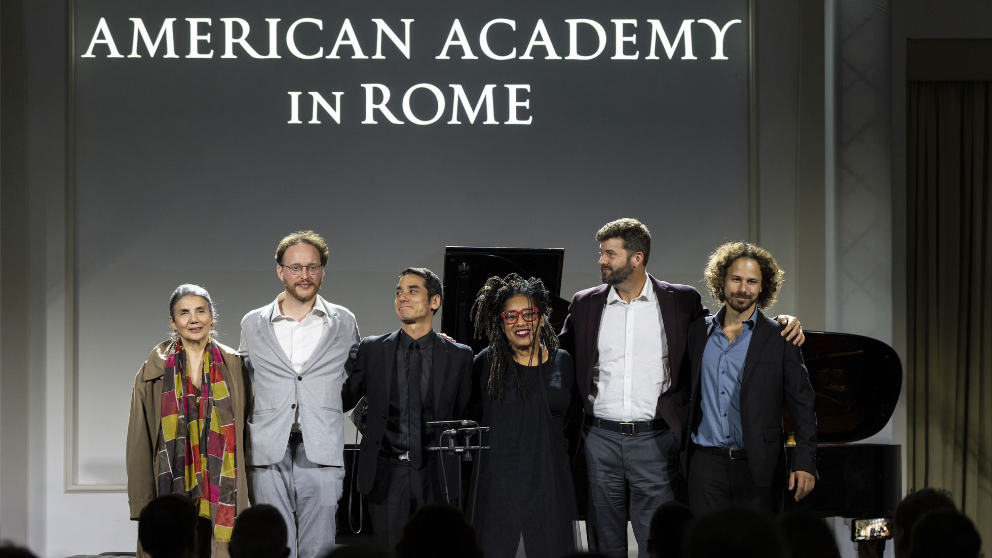 Color photograph of four men and two women standing arm and arm on a stage for musical performances