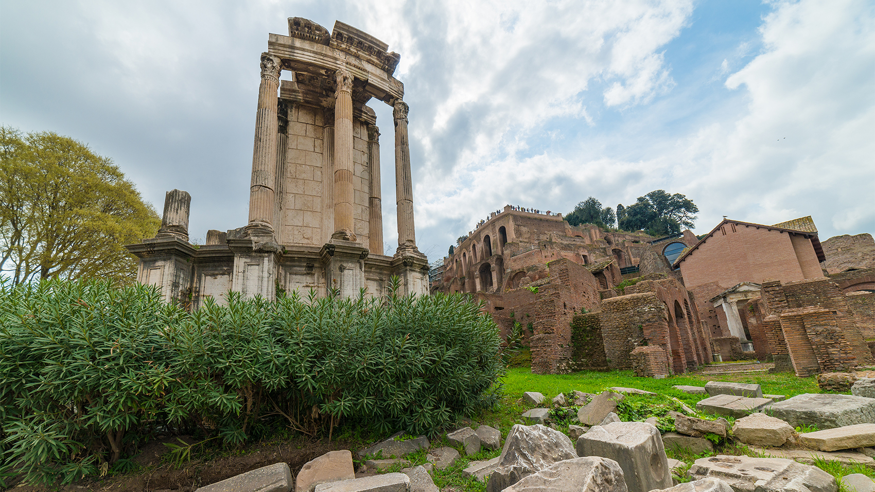 Color photograph of the ruins of an ancient Roman temple, with a narrow wall and several columns, behind a rock strewn grassy area with shrubs