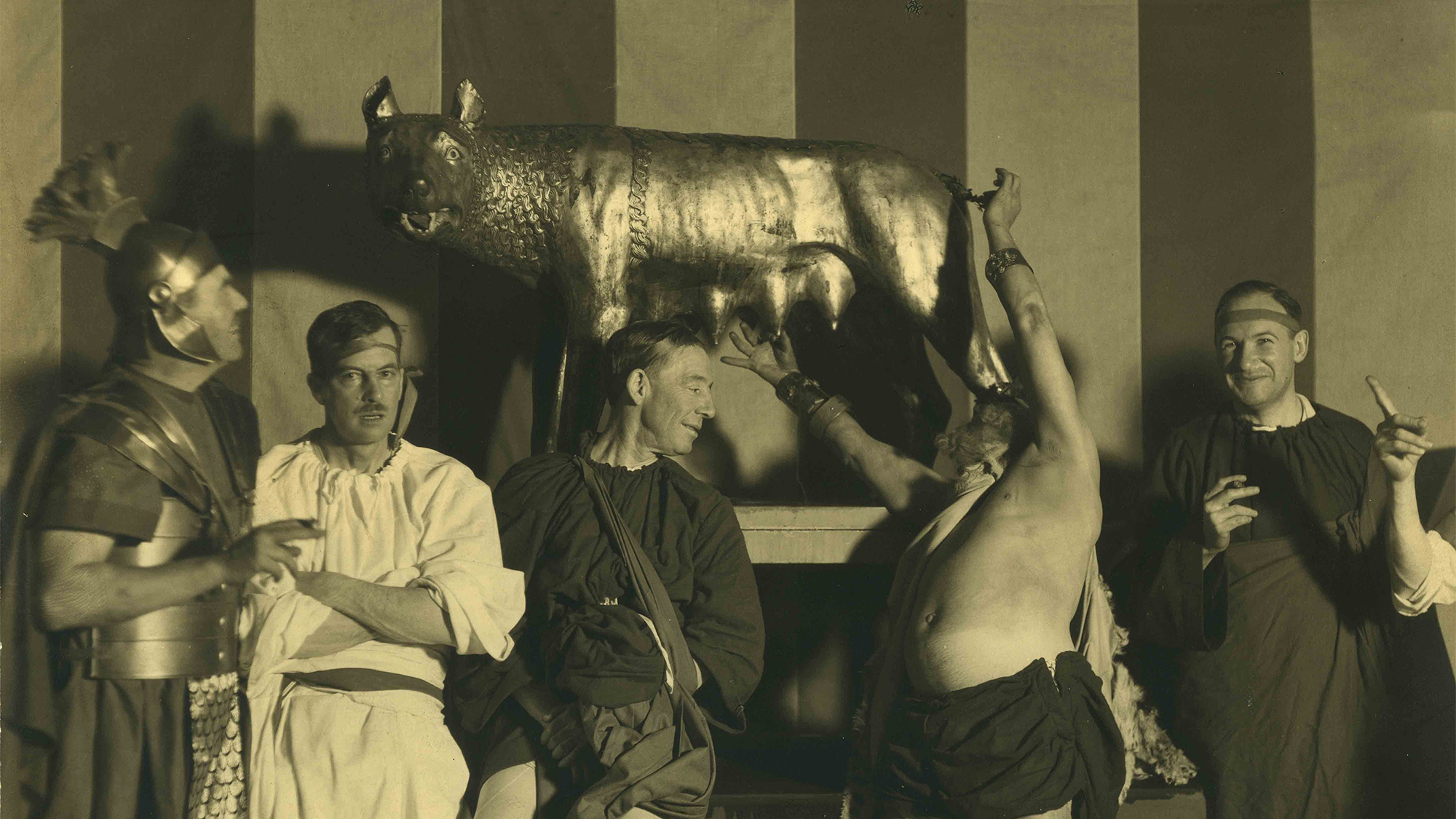 Vintage photo of men in ancient Roman costumes partying in front of a bronze she wolf statue standing on a raised platform