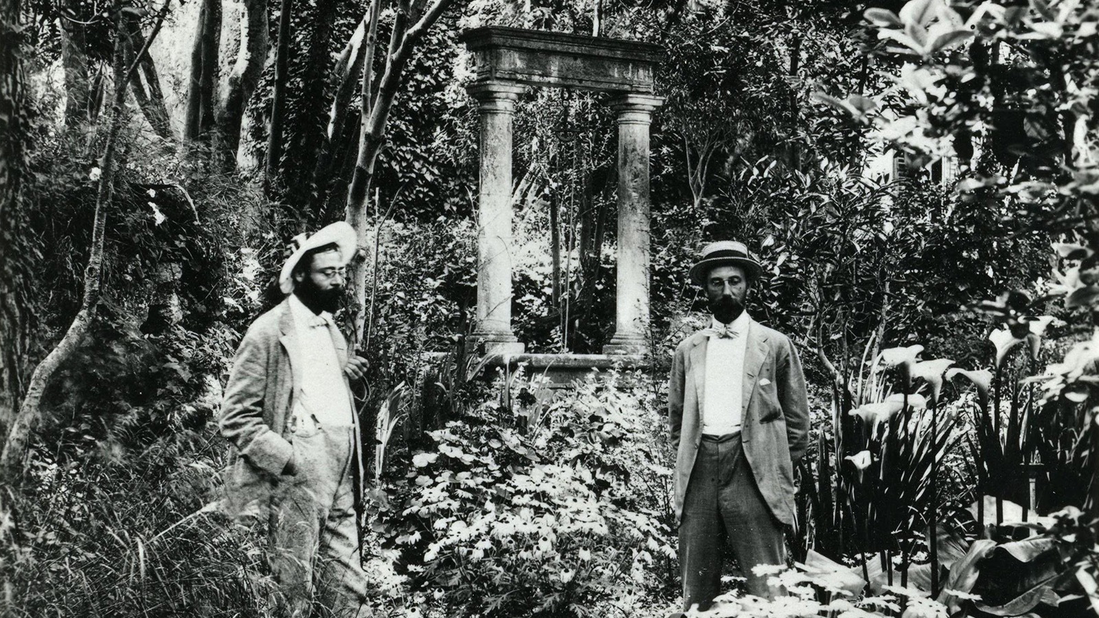 black and white photo of two men standing in a lush garden; the photo was taken over 100 years ago