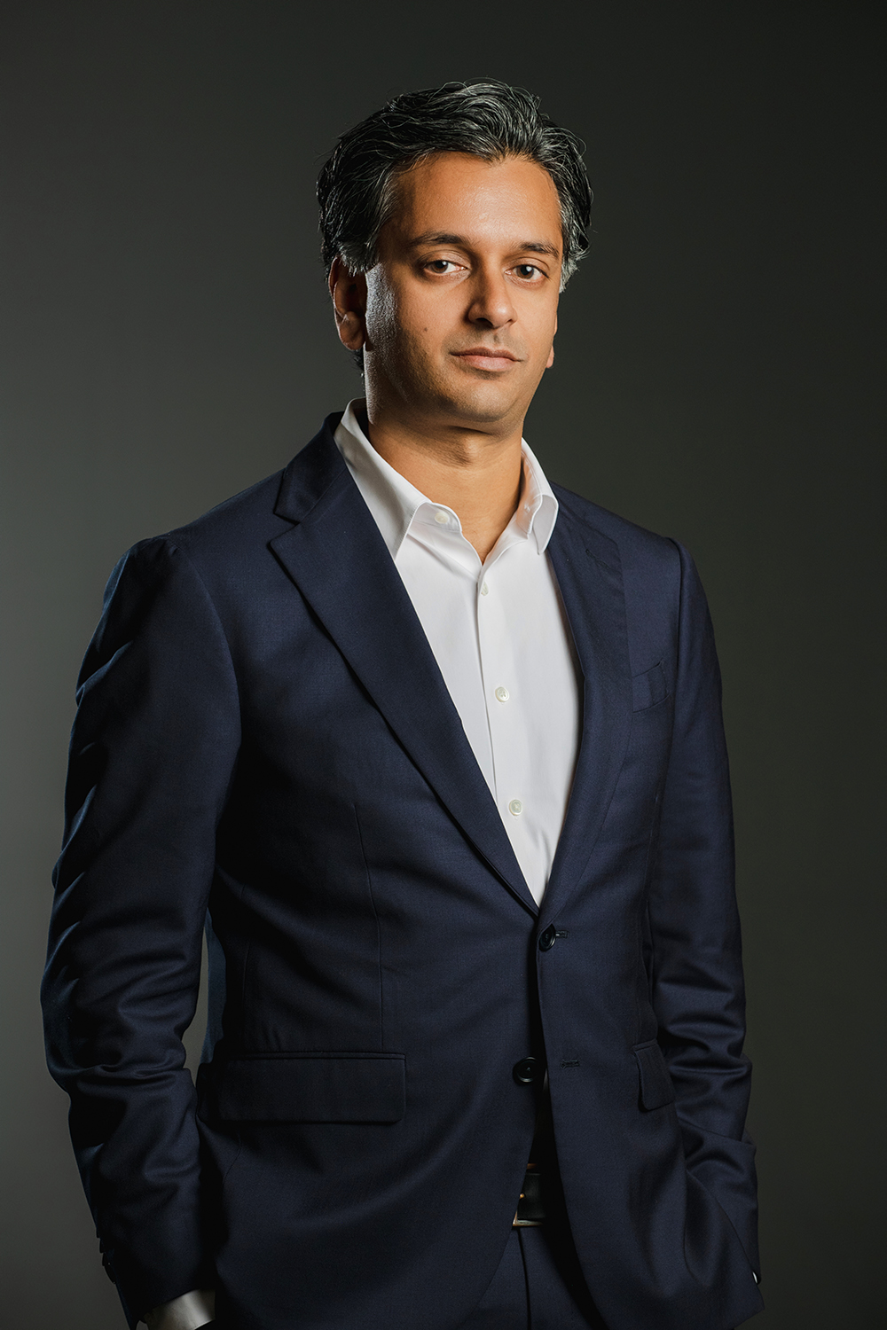 Color photograph of an olive skinned man wearing a collared shirt and dark blazer looking at the camera with a serious expression