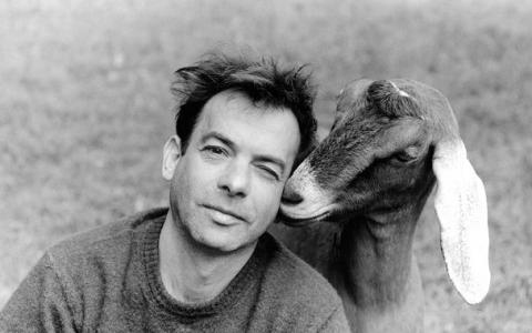 Brad Kessler Black and white photograph of the head and shoulders of a light skinned man sitting on the ground, with a goat sniffing his face