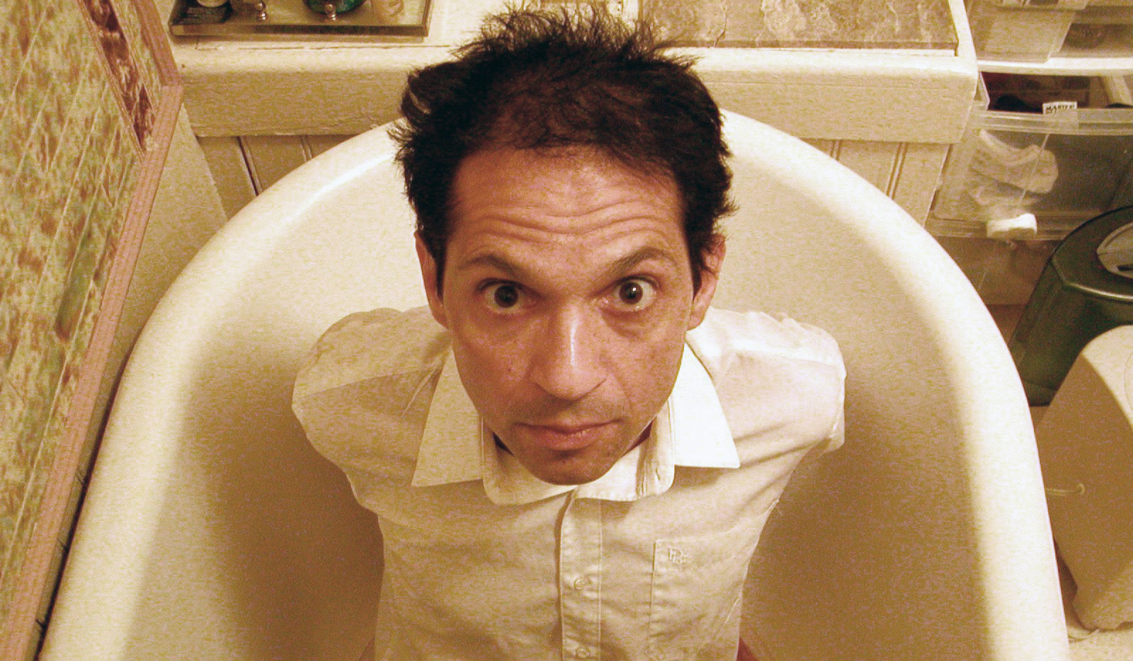 Caveh Zahedi Color photograph of the head and torso of an olive skinned man sitting in a bathtub while wearing clothes
