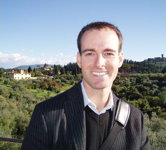 Daniel R. McReynolds Color photograph of the bust of a light skinned man smiling at the camera as he stands in a hilly green outdoor location on a sunny day