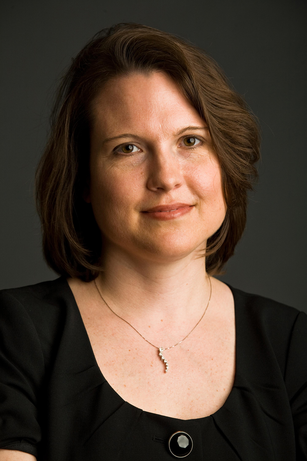 Elizabeth McCahill Color photograph of the head and torso of a light skinned woman with brown hair in a photographer's studio wearing a black top and smiling at the camera