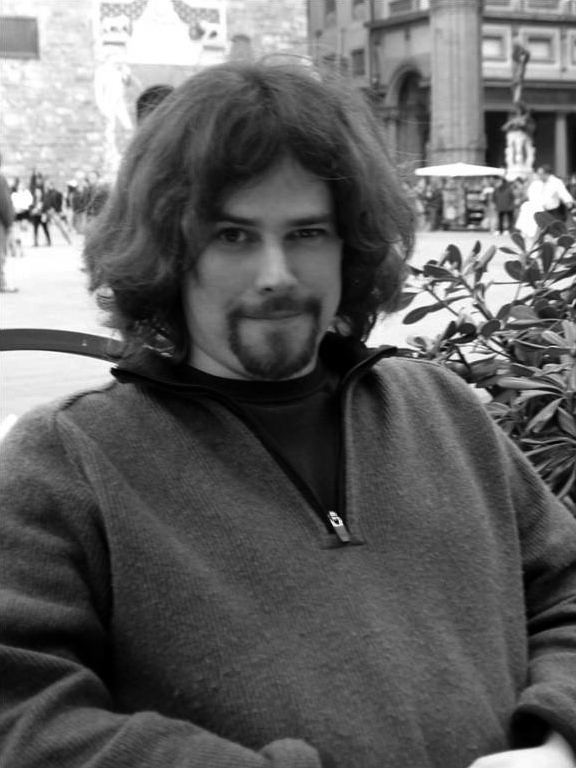 Erik Gustafson Black and white photograph of the torso of a light skinned man with long brown hair and goatee sitting in a piazza in Florence; he looks at the camera