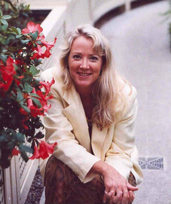 Florence Eliza Glaze Color photograph of light skinned woman wearing a cream blazer and crouching next to a planter with red azaleas