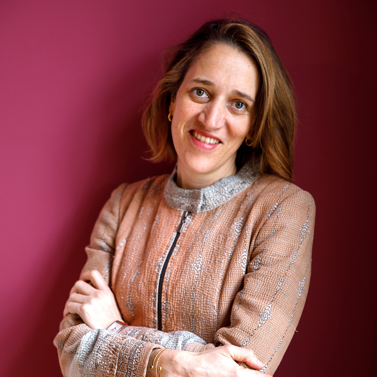 Ilaria Puri Purini Color photograph of a light skinned woman standing with her arms crossed and smiling at the camera in front of a fuchsia wall