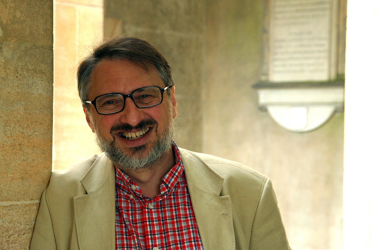 Jaś Elsner Color photograph of a light skinned man wearing a light tan blazer and red button up shirt leaning against a wall; he smiles at the camera