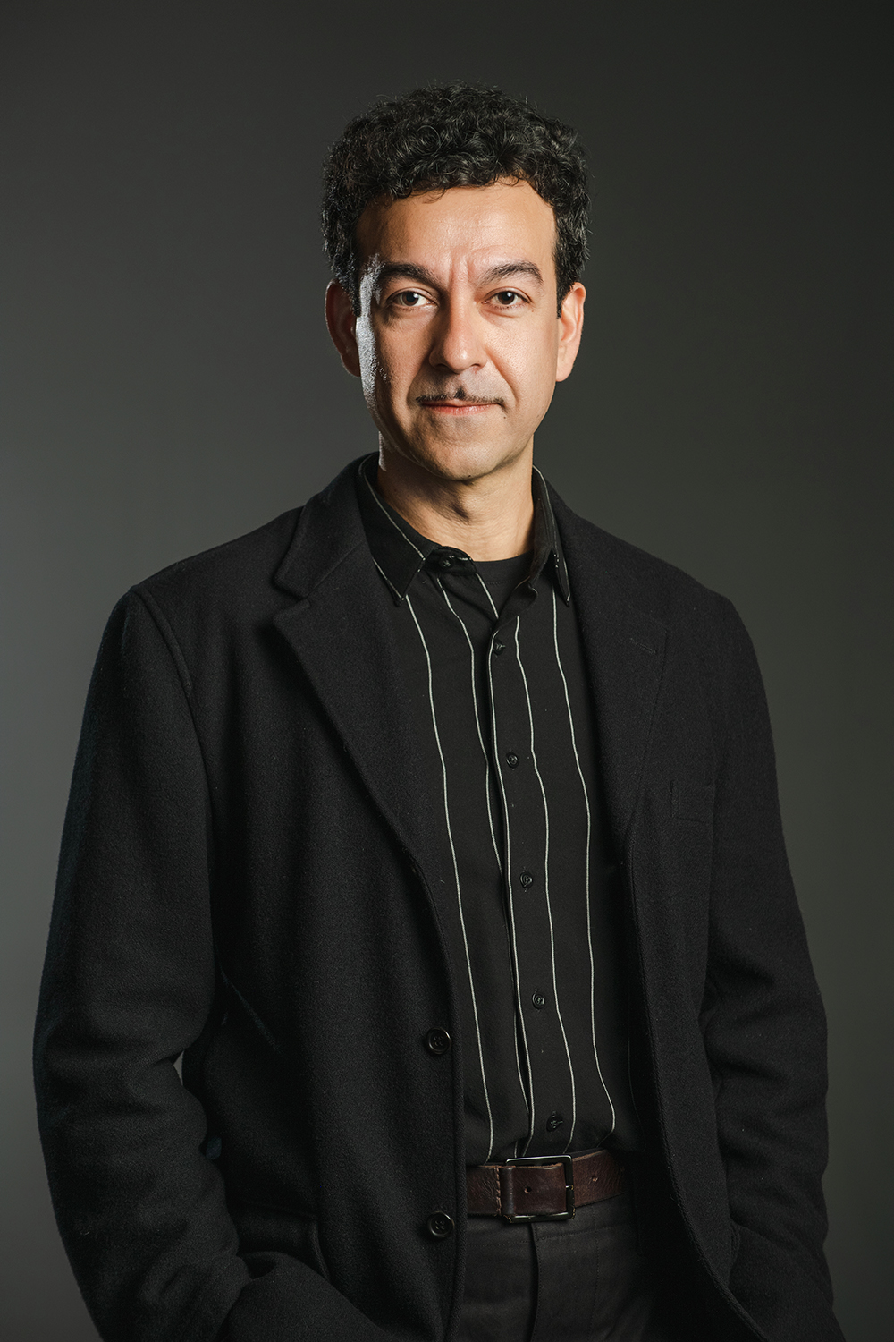 Kamrooz Aram Color photograph of an olive skinned man wearing a black blazer, shirt, and pants looking directly at the camera