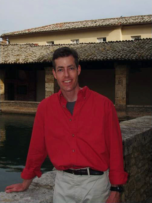Matthew Bronski Color photograph of a light skinned man from the waist up; he wears a red long sleeved shirt