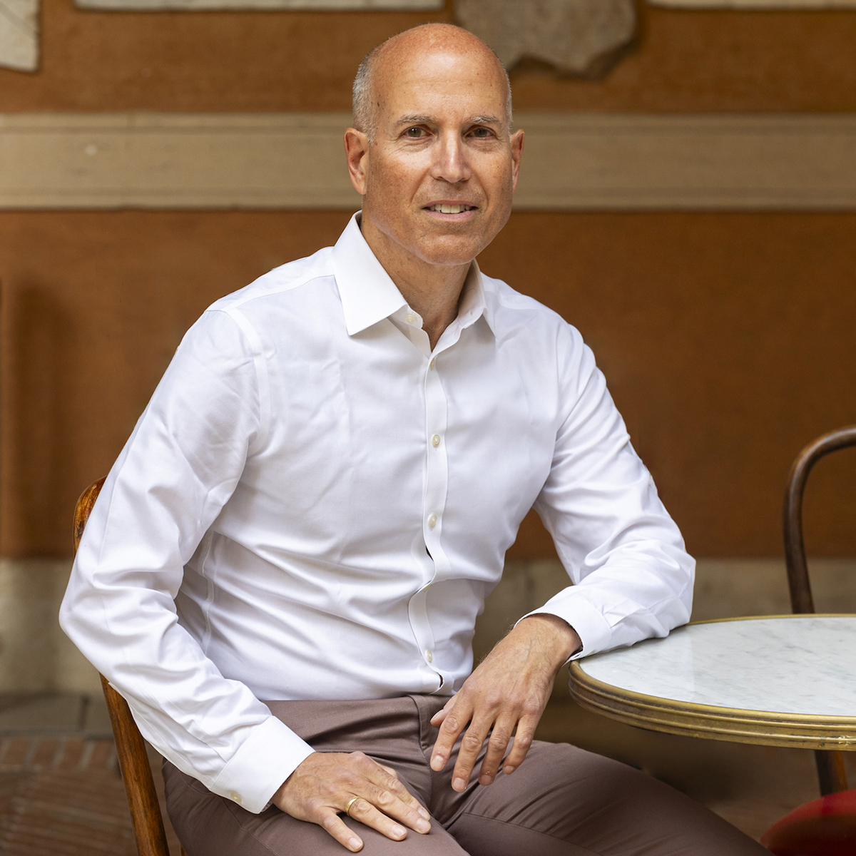 Peter N. Miller Color photograph of a light skinned man wearing a white shirt and sitting at a cafe table, smiling at the camera