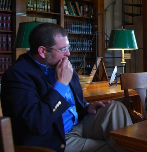 Scott Craver - library Color photograph of a light skinned man sitting in a chair in a library with a lamp and books on wooden shelves behind him