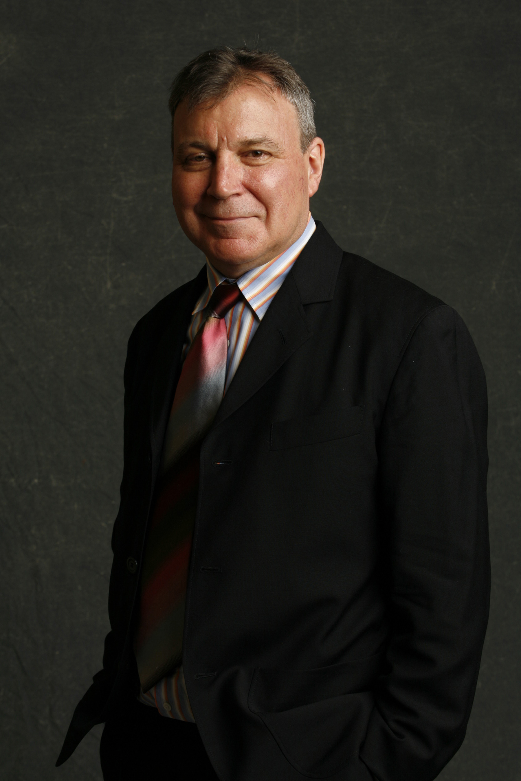 Stephen Westfall Color photograph of a light skinned man seen from the waist up in a photographer's studio wearing a suit and tie and smiling at the camera