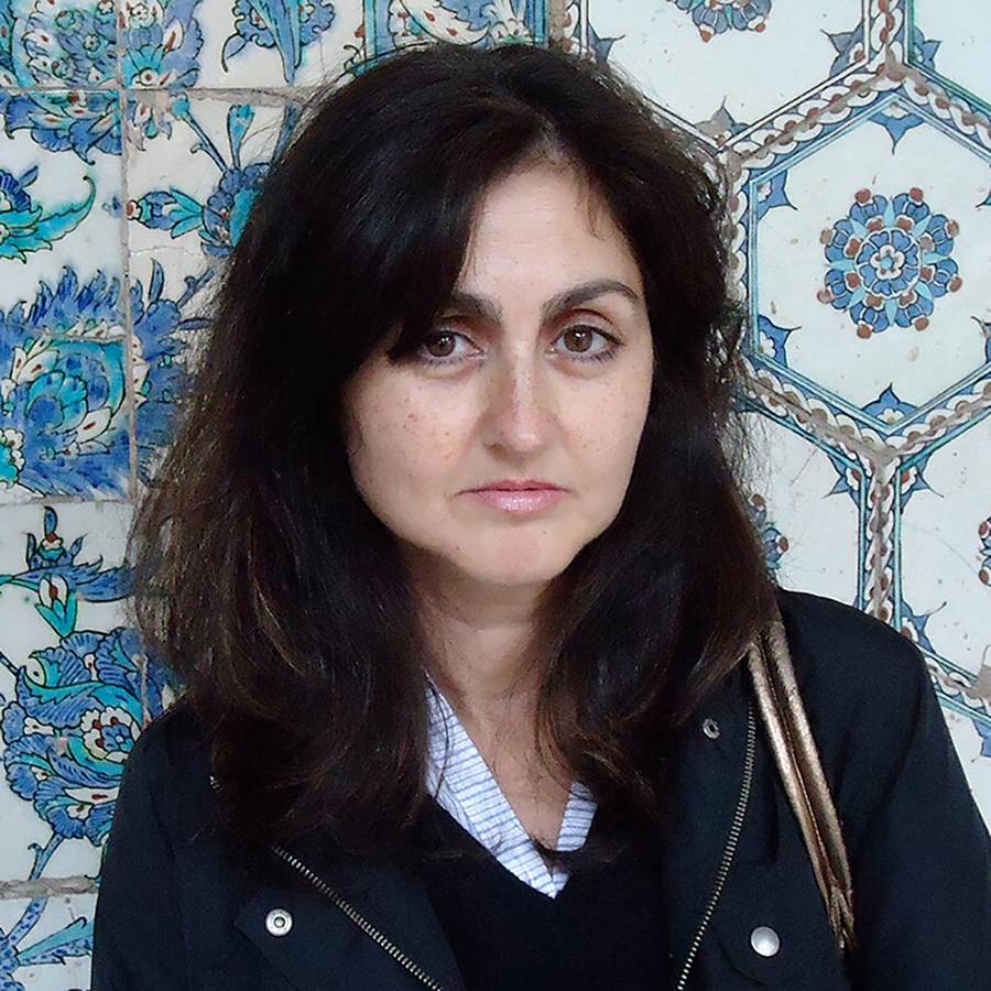 Color photograph of the head and shoulders of Zainab Bahrani standing in front a wall of painted Islamic tiles