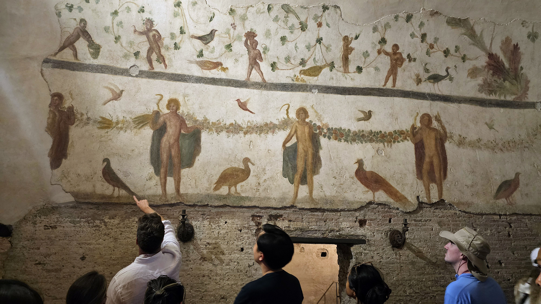 Color photo of a school group visiting an ancient roman mural, the group leader raises an arm directing attention to the wall painting