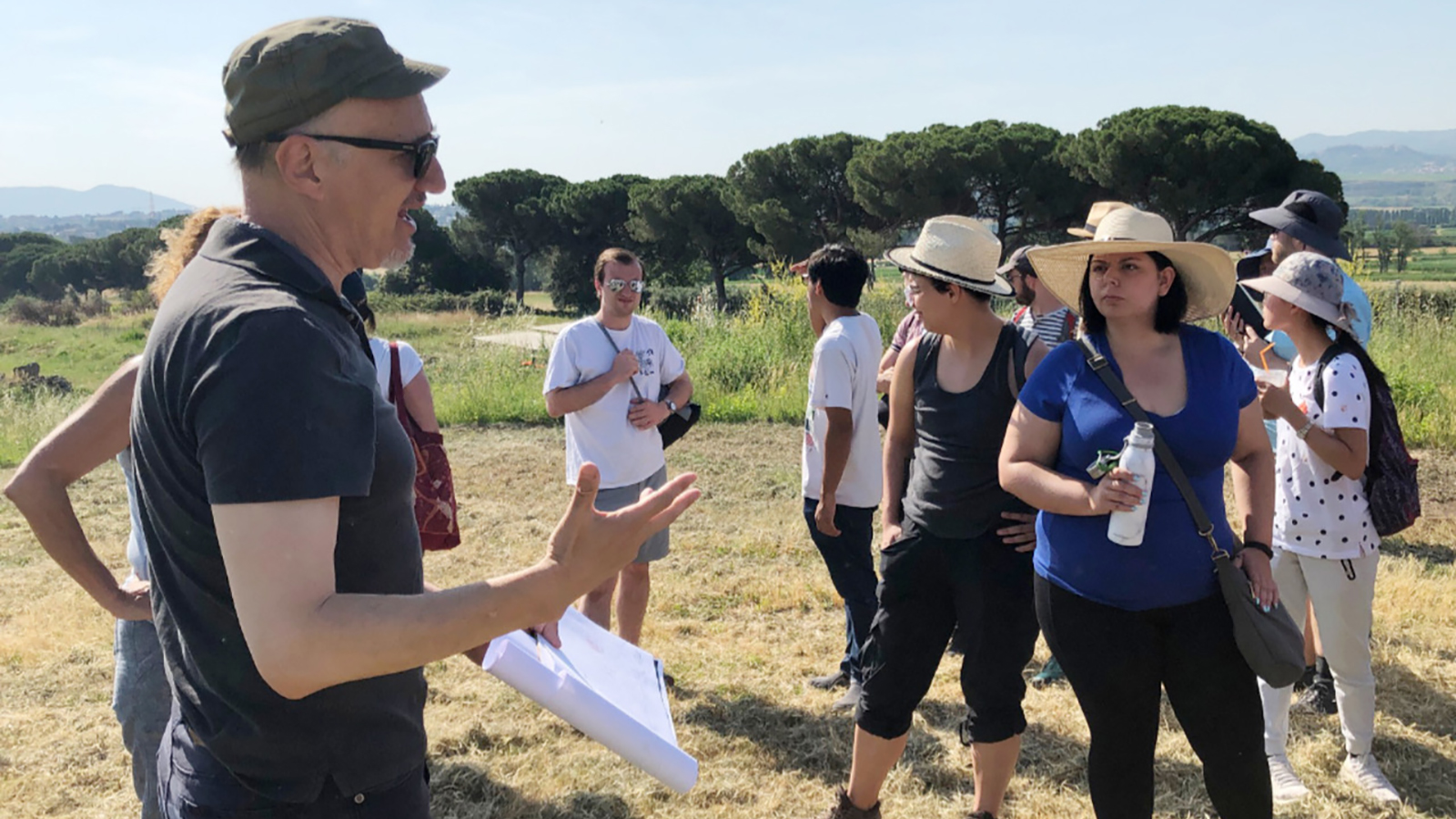 A man on the left speaks to a group of people on the right in an outdoor location at Gabii, Italy