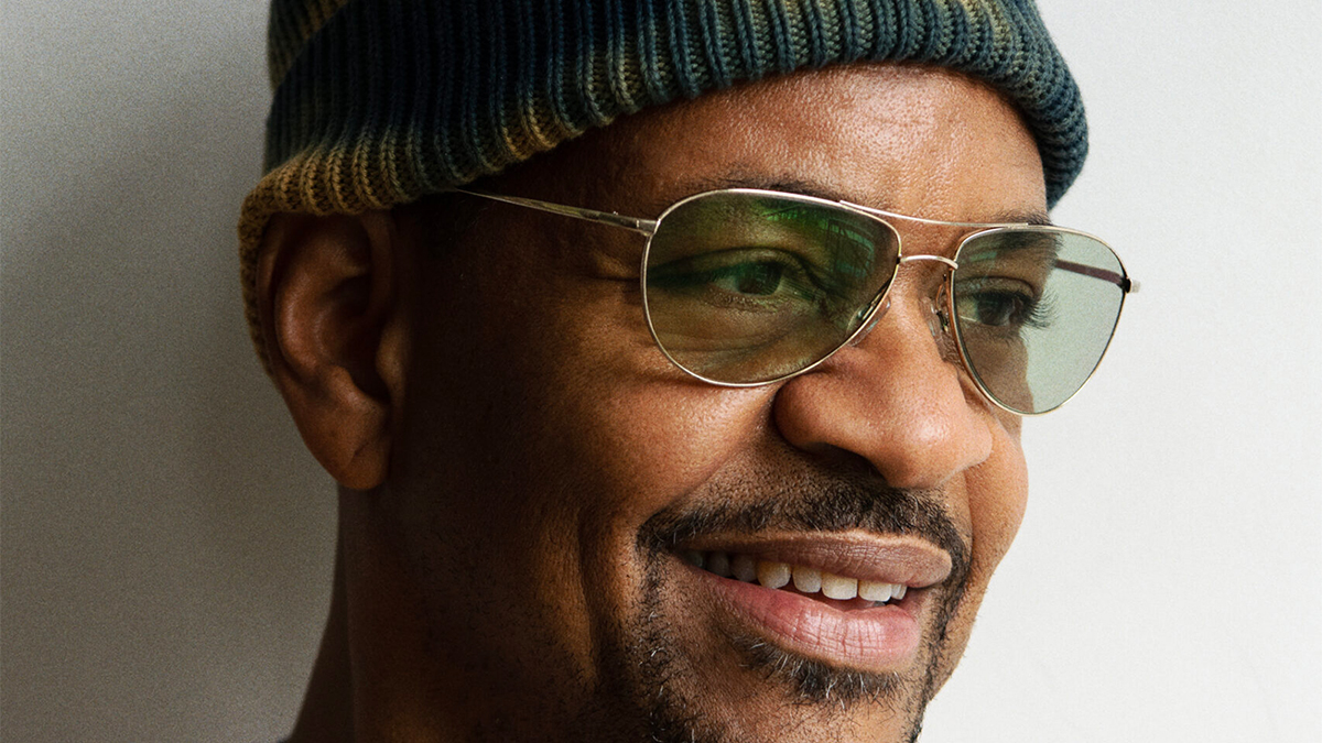 Color photo of the face of a brown skinned man wearing glasses and a hat looking off camera and smiling slightly
