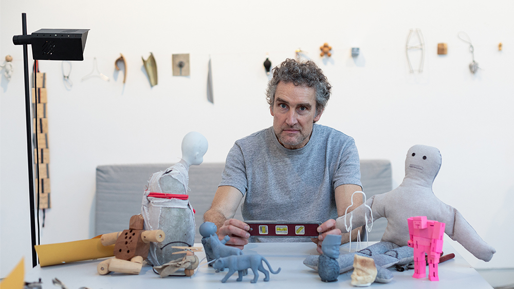 Color photo of a light skinned man sitting at a table with various small toys, looking at the camera