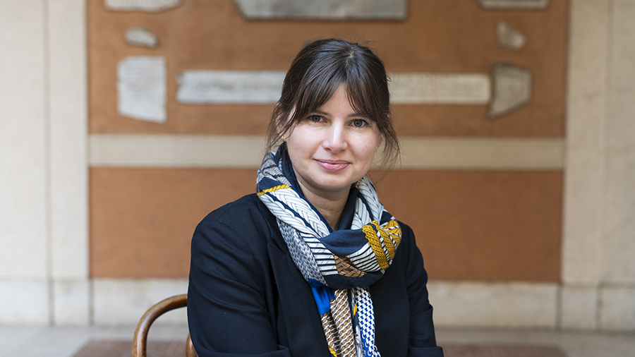 Color photo of the head and torso of a light skinned woman sitting in a courtyard and looking at the camera