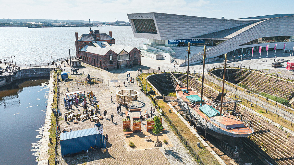 Aerial view of Canning Dock in Liverpool, showing an old sailing vessel at dock,  the Museum of Liverpool building, and various outdoor sculptures and installations alongside the river