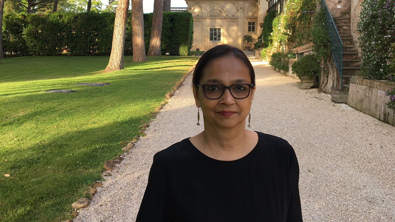Color photograph of a brown skinned woman wearing glasses and a black shirt and standing in the gardens of the Academy's Villa Aurelia; she looks at the camera with a slight smile