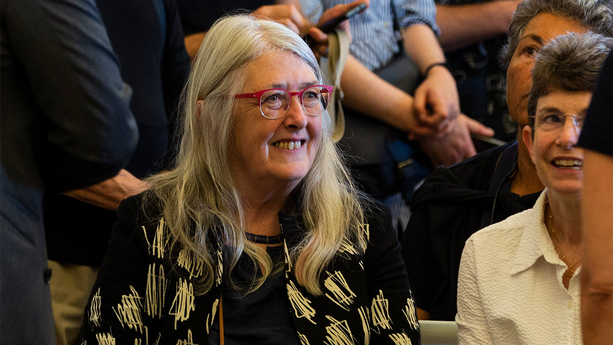 Color photograph of an older, light skinned woman with gray hair smiling and looking off camera as she sits with the audience in a lecture room