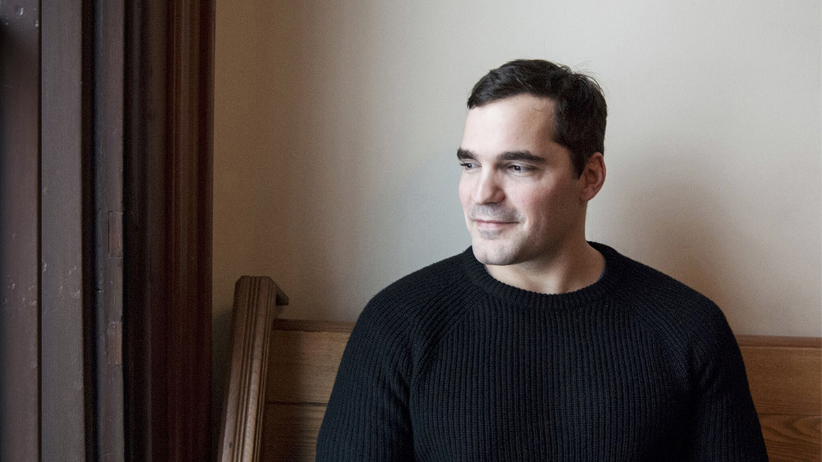 Color photograph of the head and torso of a light skinned man seated indoors on a wooden bench