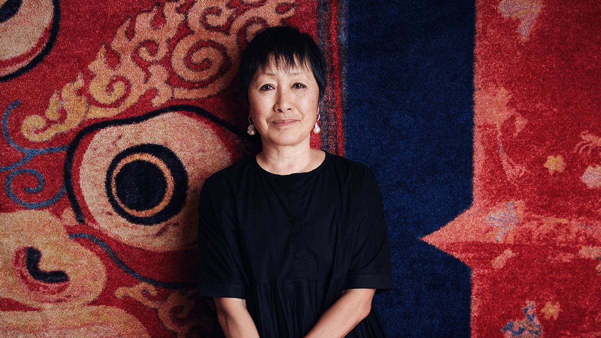 Color photograph of the head and torso of Billie Tsien, wearing a black top and standing in front of an abstract tapestry in red, blue, and black