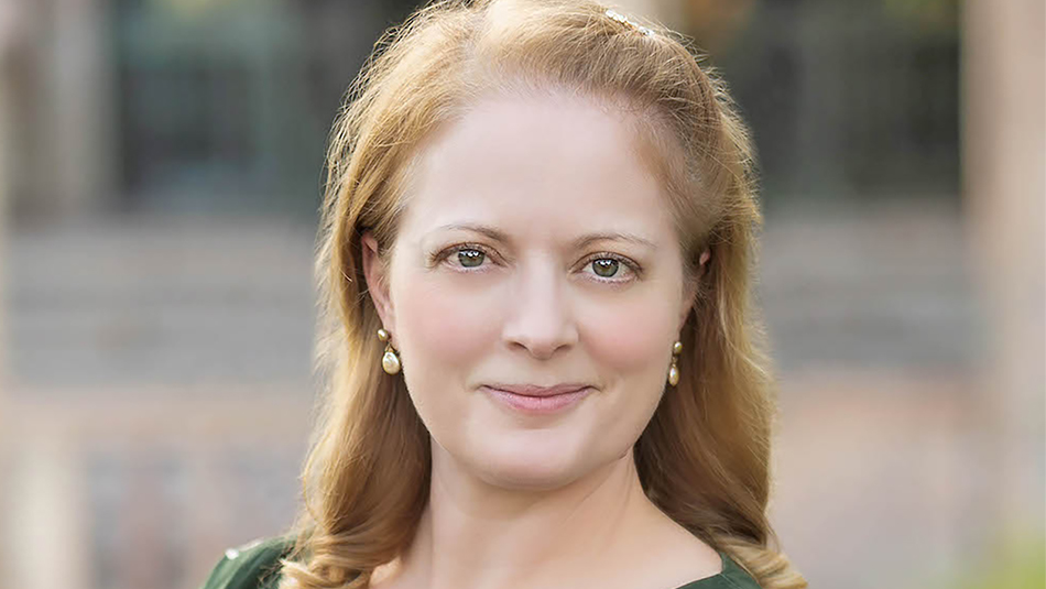 Color photograph of the head and face of a light skinned woman with strawberry blond hair smiling slightly at the camera