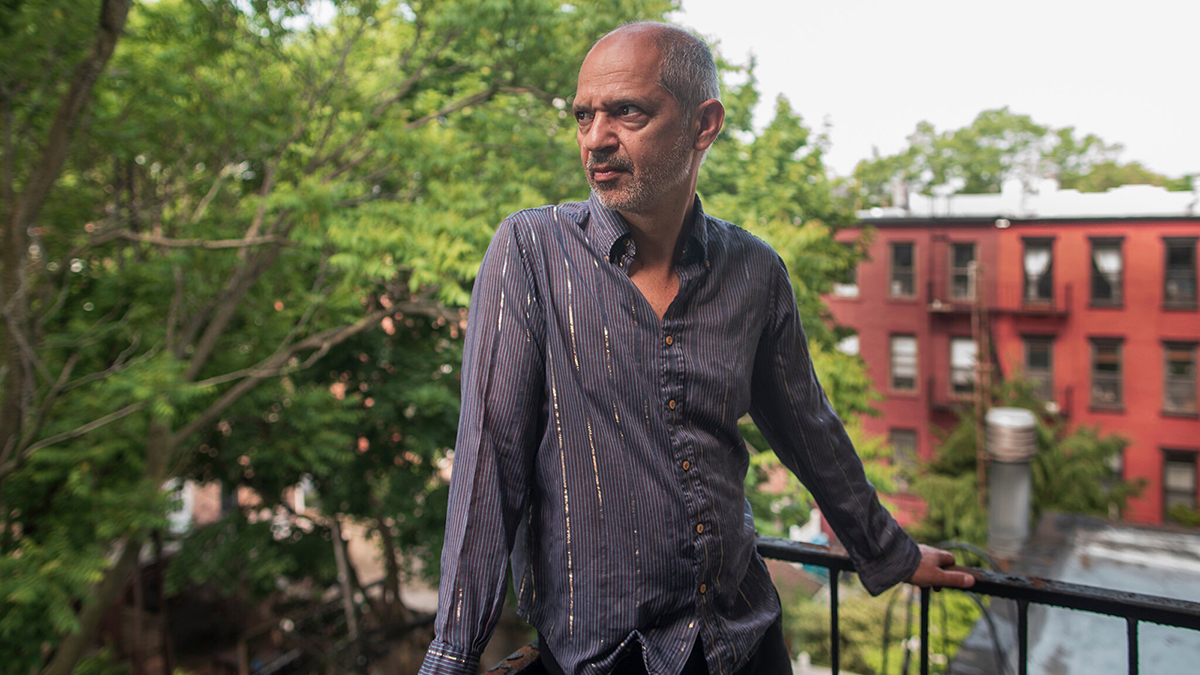 Color photograph of Caveh Zahedi standing on the outdoor backyard patio balcony of a New York apartment, overlooking the interior of the block