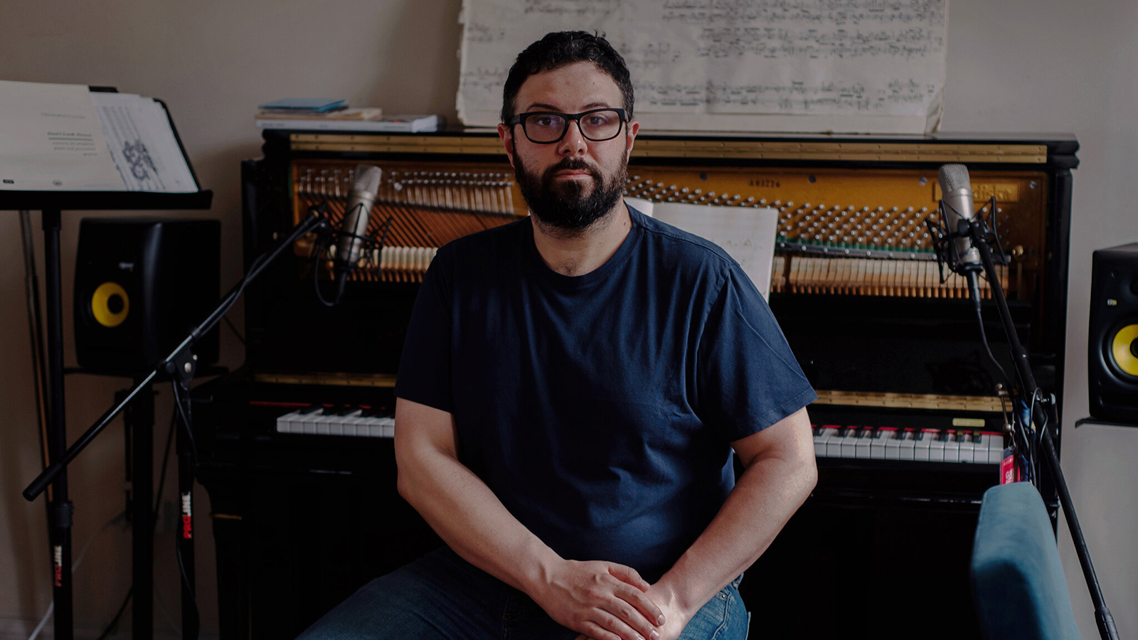 Color photograph of Christopher Cerrone in his Brooklyn apartment sitting in a piano bench with the instrument behind him