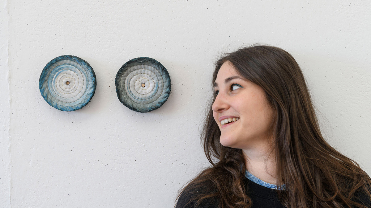 Color photograph of the head and shoulders of Corinna Gosmaro in profile, standing against a white studio wall and looking left at two blue and white circular fabric artworks on the wall, hanging at eye level