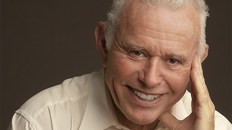 Color photo of the head and shoulders of an older light skinned man, smiling at the camera as his hand touches his cheekbone