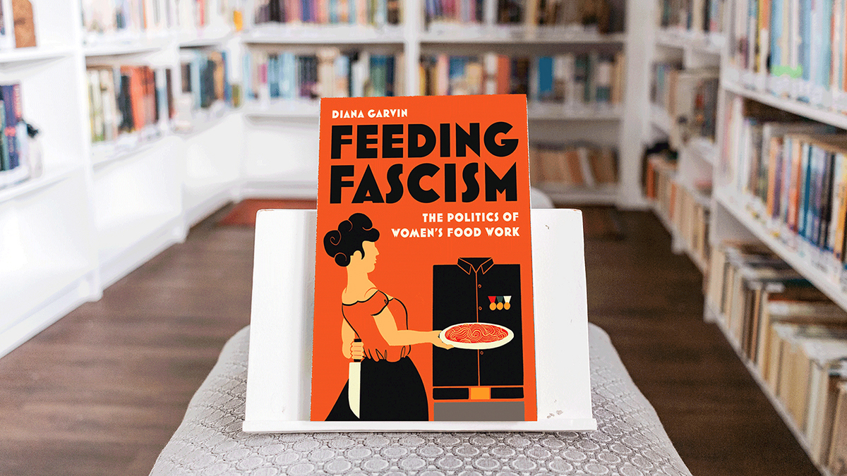 Color photograph of Diana Garvin's book Feeding Fascism leaning against a wooden support and propped on a cushion, in a room with three walls that contain bookshelves full of books