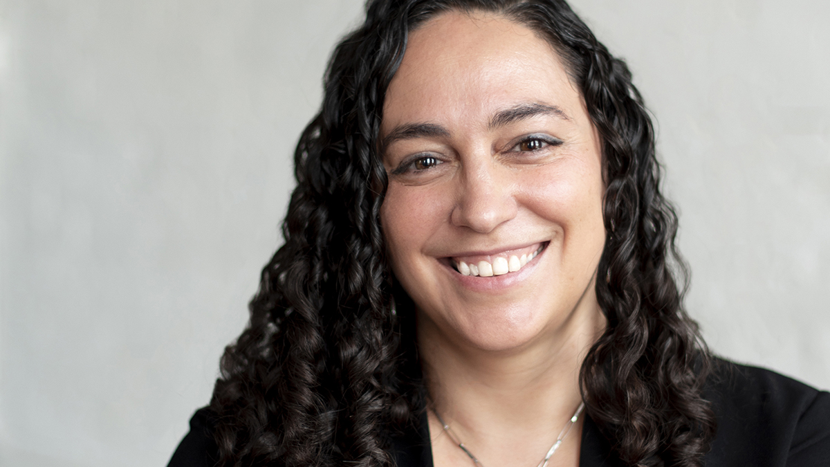 Color photograph of the head and face of a light skinned woman with curly brown hair who smiles at the camera