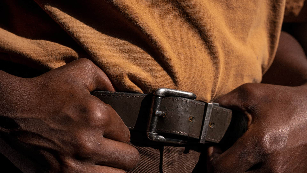 Detail of a color photograph of the midsection of a human torso; a person wearing a yellow shirt digs their hands into a brown belt