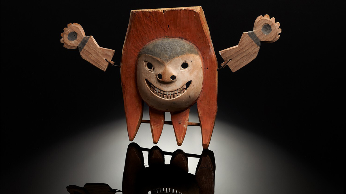 Color photograph of an indigenous Alaskan carved wood mask that depicts a face with outstretched arms